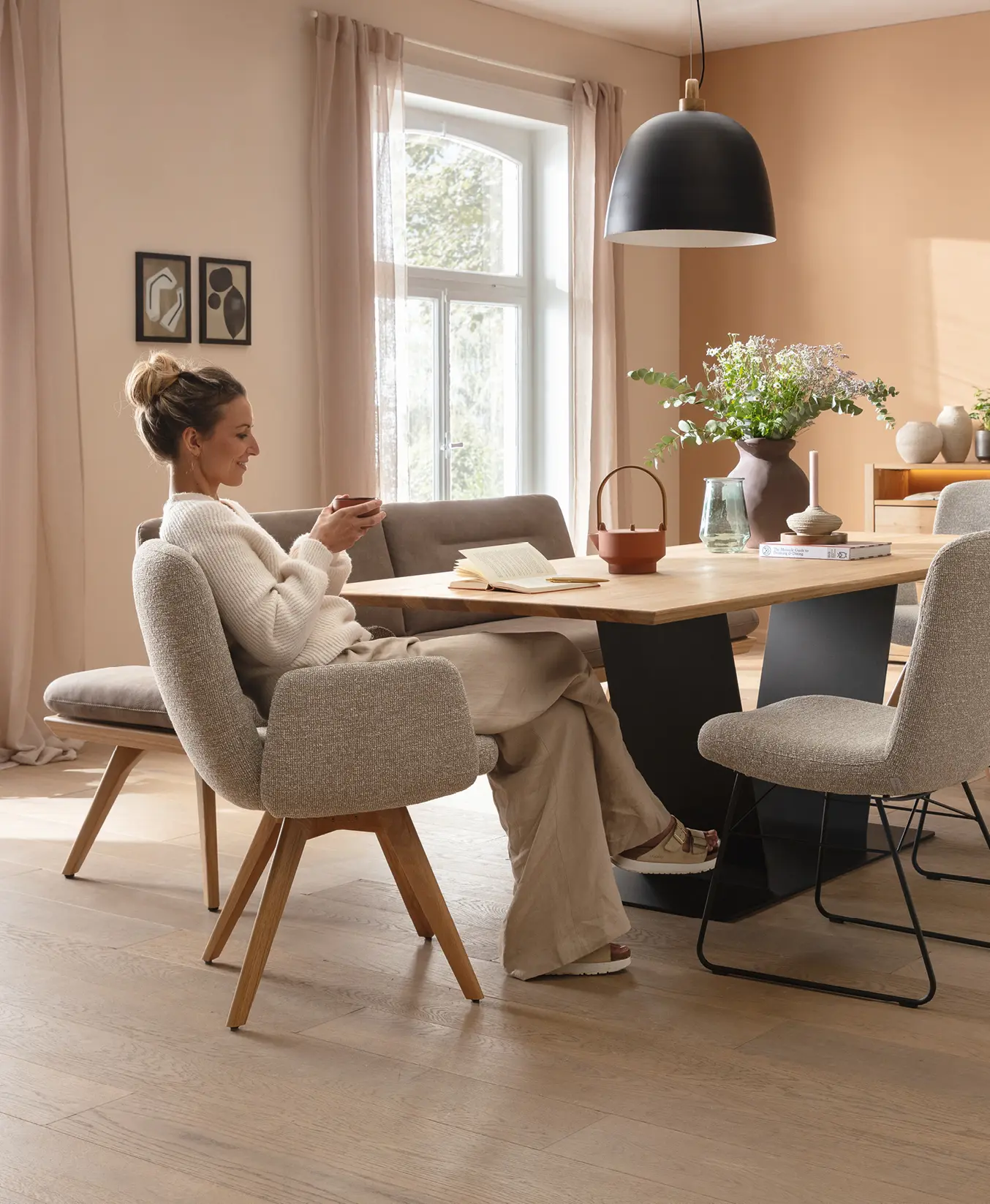 Frau sitzt am Esstisch mit gepolsterten Stühlen, Buch und Deko auf der Tischplatte, Fenster und Bilder an der Wand